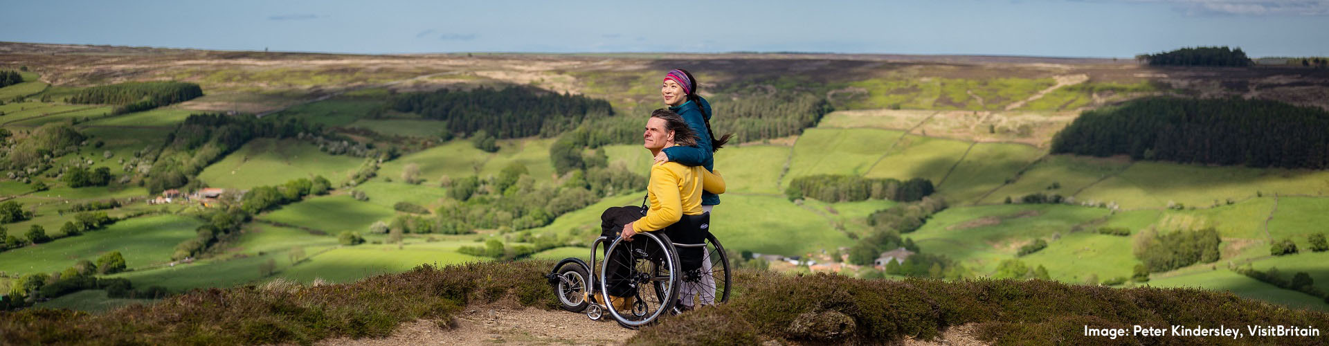 A distanced perspective of two people (a male in a manual wheelchair and a femme stood next to him) arm in arm at the top of Bank Top, Rosedale, overlooking a valley of fields and trees.